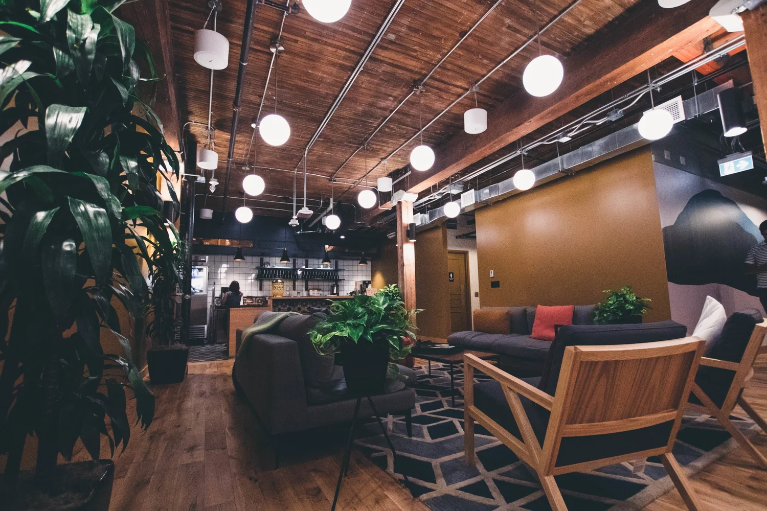 Grande salle de réunion moderne avec canapés, fauteuils en bois, plantes d’intérieur et éclairage suspendu à spots ronds.